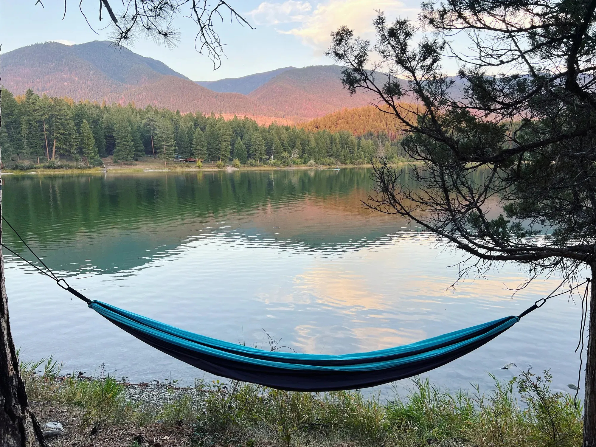 A hammock strung between two trees at the edge of a still lake, with forested hills and mountains behind it at sunset.
