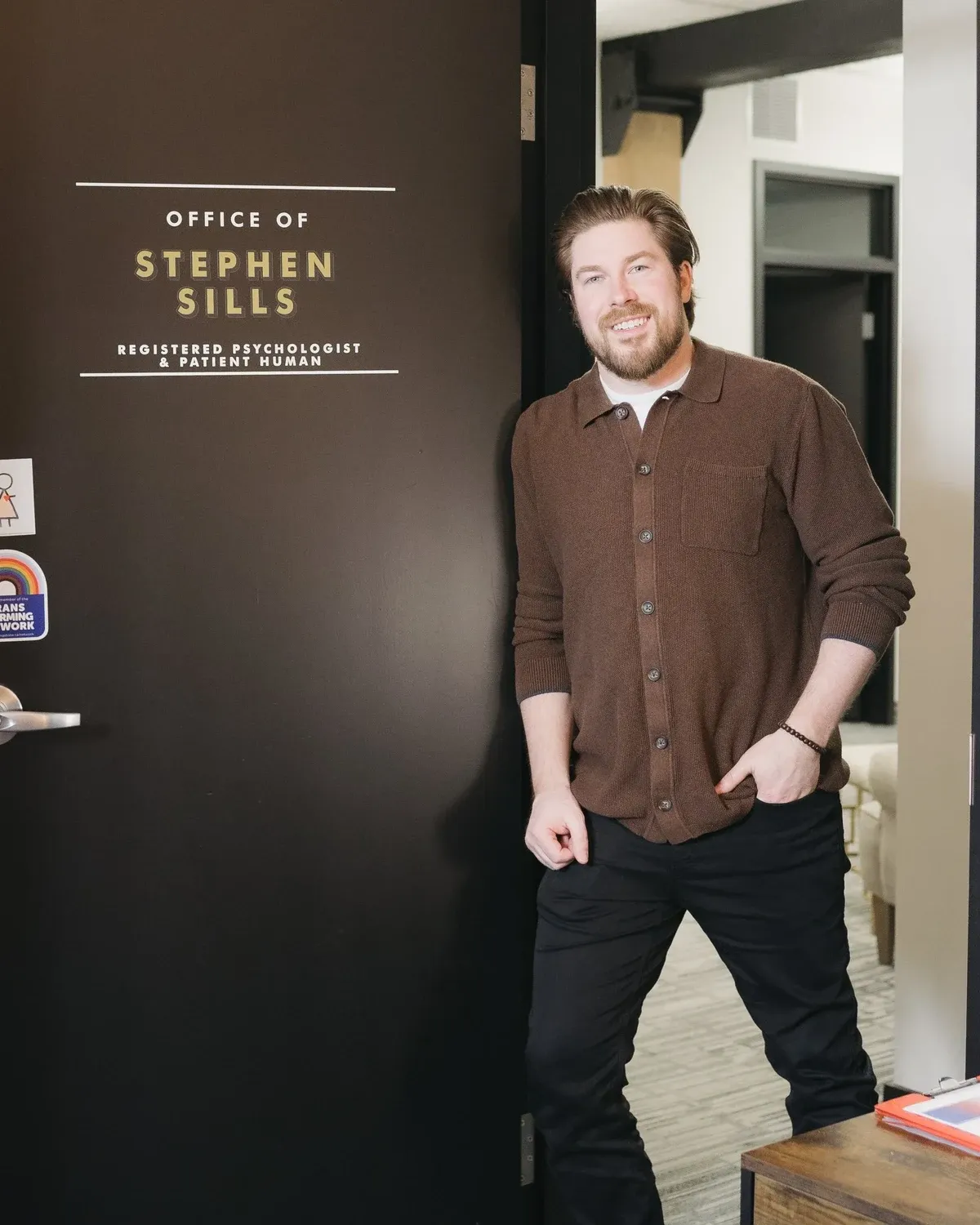 Stephen Sills smiling in the doorway of his office. The door reads: Office of Stephen Sills, Registered Psychologist and Patient Human.